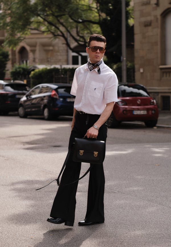 Camicia bianca a maniche corte, pantaloni larghi neri, borsa in pelle nera con accessori in oro, occhiali da sole e sciarpa a fantasia. Sfondo urbano.
