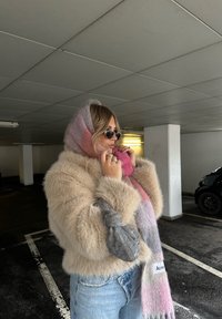 Beige nepbontjas met een roze en grijs ombre-sjaal, grijze textuur tas en ronde zonnebril, gezet in een parkeergarage.