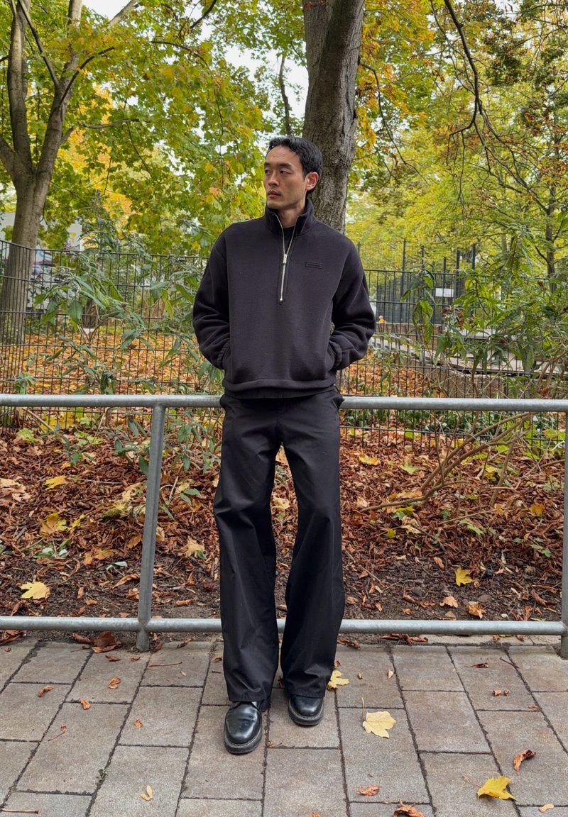 Un homme en pull foncé et pantalon se tient près d’une rambarde dans un parc automnal, les mains dans les poches et le regard tourné sur le côté.