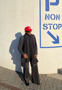 Μαύρο κομμένο παλτό πάνω από μια μαύρη μπλούζα, φαρδύ παντελόνι και κόκκινο καπέλο με λογότυπο. Το φόντο έχει ένα λευκό τοίχο με μπλε κείμενο.