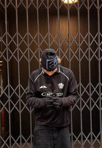 Czarna koszula z długim rękawem z białymi akcentami, czarna czapka z logo, czarne rękawiczki; osoba stoi z rękami złożonymi przed metalową bramą.