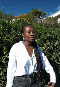 Una persona che indossa una camicia bianca con bottoni e jeans neri a vita alta si trova di fronte a una vegetazione verde. Orecchini d'argento notevoli accentuano il look.