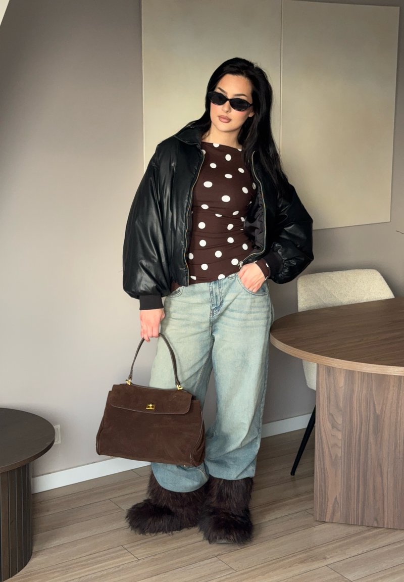 Femme portant une veste matelassée noire, un haut marron à pois, un jean ample, des bottes en fourrure, des lunettes de soleil, tenant un grand sac à main marron à l'intérieur près d'une table en bois.