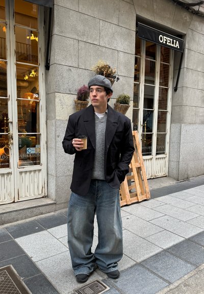Blazer negro sobre un suéter gris, jeans de mezclilla azul de corte holgado, boina gris, y sosteniendo un vaso de café desechable, de pie en una calle de la ciudad.