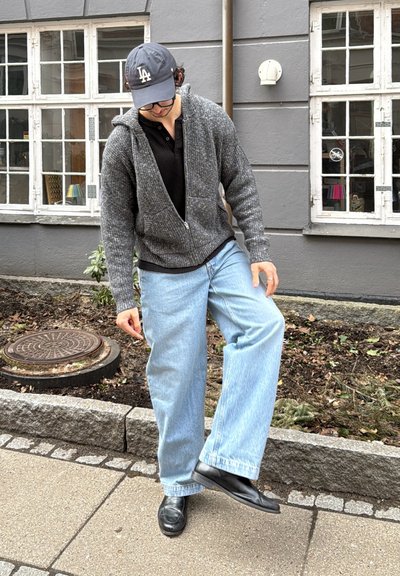 Persona con una sudadera gris con capucha, camiseta negra, vaqueros azul claro, zapatos negros y una gorra oscura que pisa una acera cerca de un edificio con ventanas blancas.