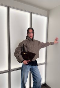 Γυναίκα με καρό σακάκι και τζιν κρατά σκούρο καφέ τσάντα, ποζάροντας με ένα χέρι τεντωμένο μπροστά σε φωτιζόμενο πάνελ τοίχο.