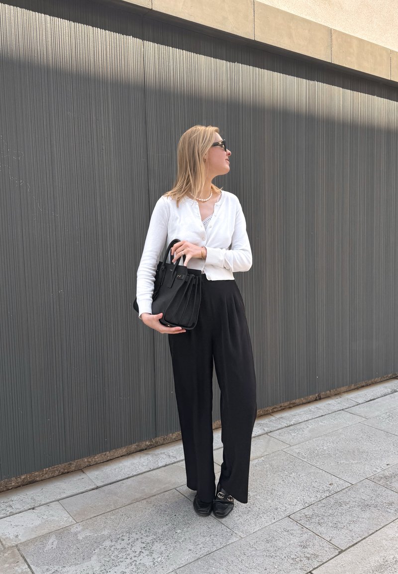 Mujer con cabello rubio que lleva gafas de sol negras, top blanco, pantalones negros y zapatos negros, sosteniendo un bolso negro, de pie contra una pared acanalada.