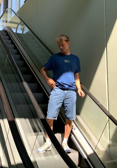 Camiseta azul de Nike con logo, pantalones cortos de mezclilla y zapatillas ligeras. Persona en una escalera mecánica, con la luz del sol creando contrastes y sombras.