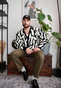 Pulover s črno-belo zebravico, olivno zeleni hlače, črne čevlje, večplastne verižice, s rastlino in umetniškim delom v ozadju.