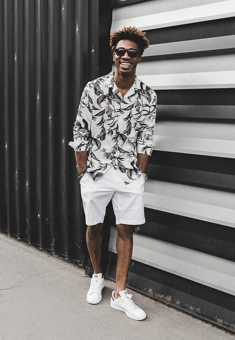 White shirt with black floral patterns, white shorts, and white sneakers. The individual stands against a striped metallic background.
