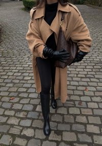 Femme marchant sur une rue pavée, portant un manteau couleur camel, des gants noirs, des leggings noirs, des bottes noires montantes et tenant un sac à main marron à motif.