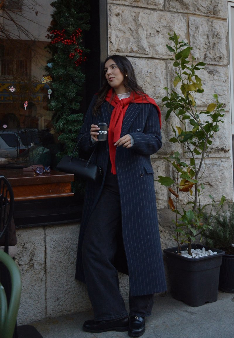 Manteau long rayé bleu marine avec une écharpe rouge, pantalon foncé et chaussures noires ; tenant une tasse de café devant un mur de pierre et de la verdure.