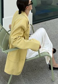 Pārlieku liels dzeltenais žakets no teksturētas auduma, kombinēts ar baltu satīna svārku. Modelis sēž uz zaļas krēsla ar melniem apaviem.