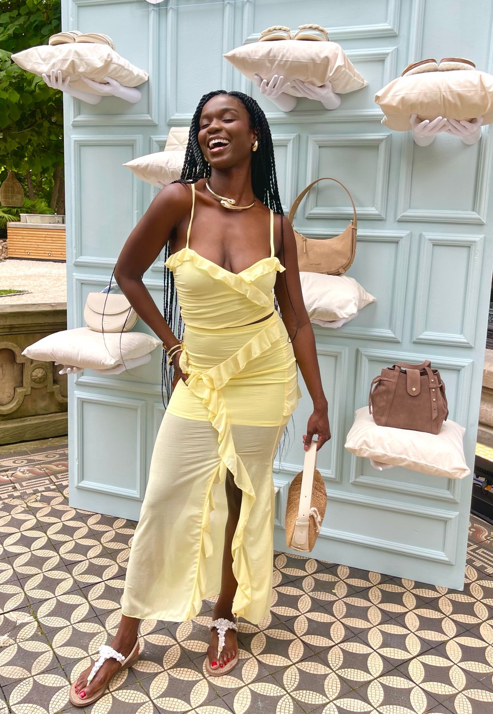 Dzeltena bez piedurknēm kleita ar viļņotām malām, izgriezumu apakšā un mīkstu audumu. Aksesuāri ietver salmu somu un sandales ar baltām detaļām.