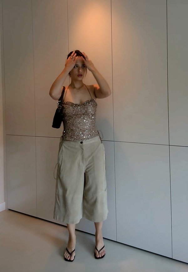 Hnedý top so flitrami a tenkými ramienkami, kombinovaný s svetlo béžovými širokými nohavicami. Čierna kabelka a žabky dopĺňajú outfit. Neutrálny pozadie.