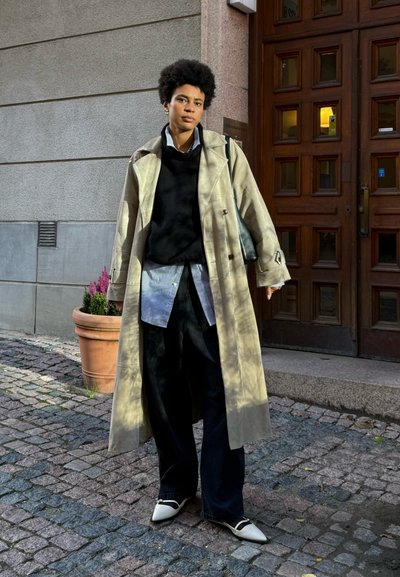 Abrigo trench beige llevado sobre un suéter negro y una camisa a rayas azul, combinado con pantalones anchos negros y zapatos blancos de punta.