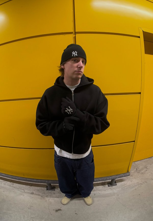 Sort zip-up hoodie, hvid t-shirt under, mørkeblå jeans, sort strikket beanie med logo, sorte handsker, stående mod en gul væg.