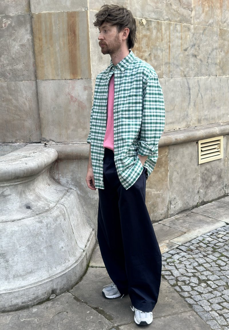 Homme aux cheveux bruns et barbe portant une chemise verte à carreaux par-dessus un t-shirt rose, un pantalon large foncé et des baskets blanches, debout près d'un mur en pierre.