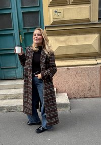 Dugi karirani kaput u tamno smeđoj, crnoj i bijeloj boji, uparen s crnim toplim i širokim plavim trapericama; drži bijelu šalicu kave.