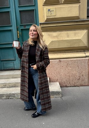 Largo abrigo de cuadros en marrón oscuro, negro y blanco, combinado con una blusa negra y pantalones vaqueros anchos azules; sosteniendo una taza de café blanca.