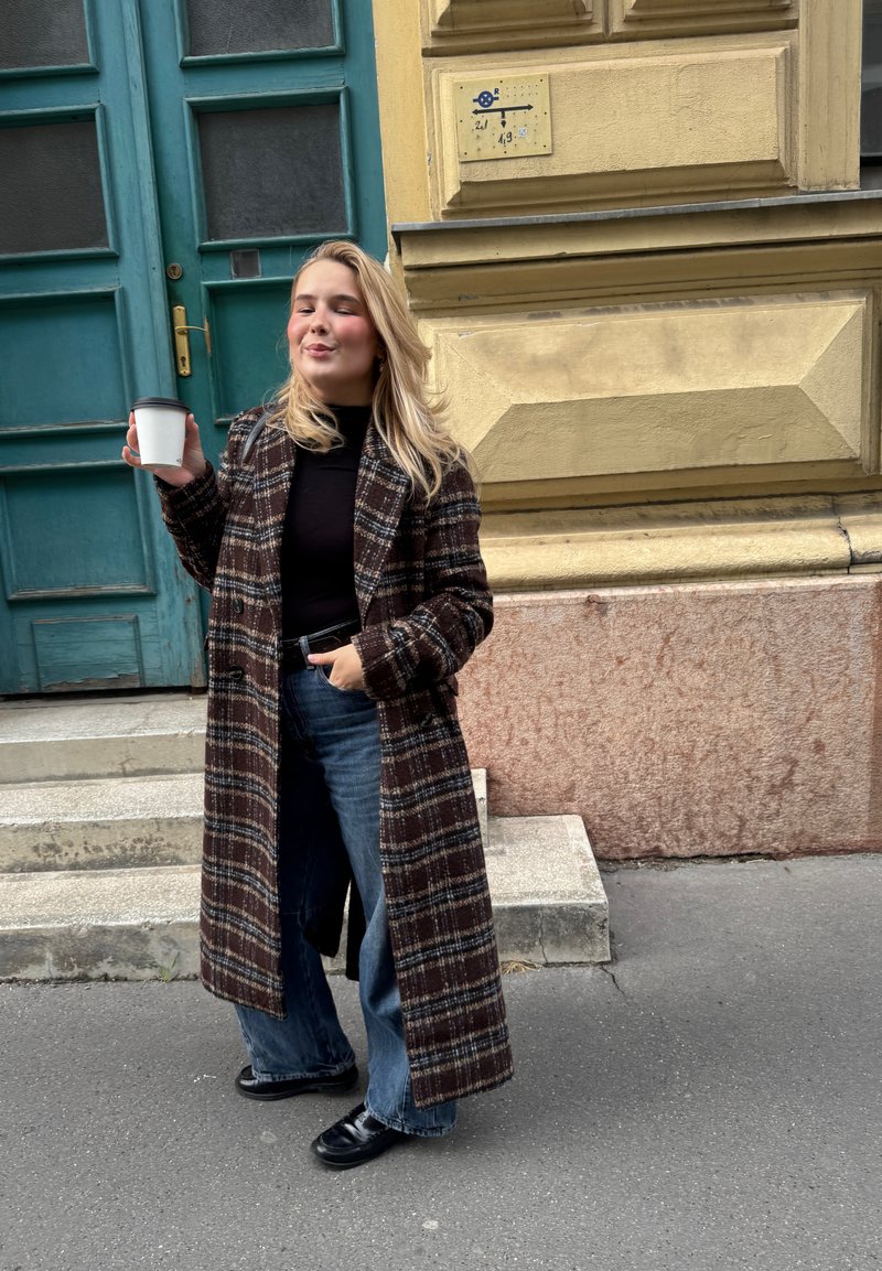 Lång rutig kappa i mörkbrunt, svart och vitt, kombinerad med en svart topp och blå vida jeans; håller i en vit kaffekopp.