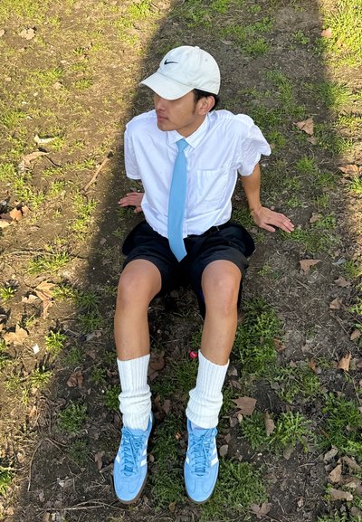 Camisa blanca de botones, corbata azul clara, pantalones cortos negros, calcetas blancas hasta la rodilla y zapatillas azules. Sentado en la hierba con una gorra puesta.