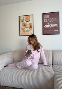 Zestaw piżam w różowo-białe paski, wykonany z miękkiej tkaniny, siedzący na beżowej sofie z prążkowanym obiciem, trzymający zielone pudełko z prezentem ozdobione świątecznymi akcentami.