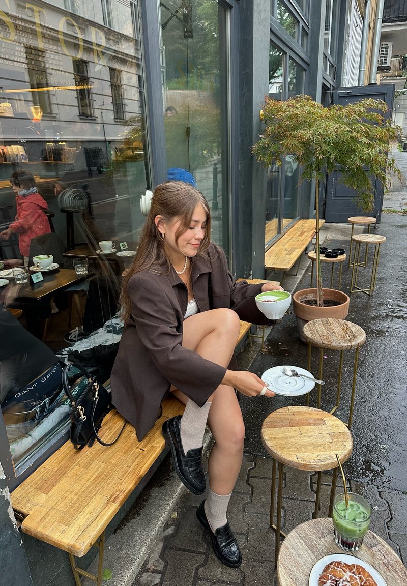 Giovane donna in giacca marrone seduta fuori da un caffè con una tazza di matcha latte, circondata da tavoli e sgabelli in legno e un piccolo albero nelle vicinanze.