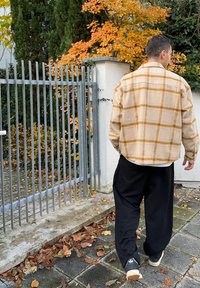 Camicia di flanella a quadri beige con linee gialle e marroni, abbinata a pantaloni neri larghi e sneakers nere, su uno sfondo di foglie autunnali.