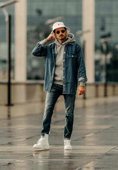 Chaqueta de mezclilla sobre una sudadera gris con capucha, jeans azules y zapatillas blancas de caña alta. Los accesorios incluyen un sombrero tipo bucket blanco y gafas de sol oscuras.