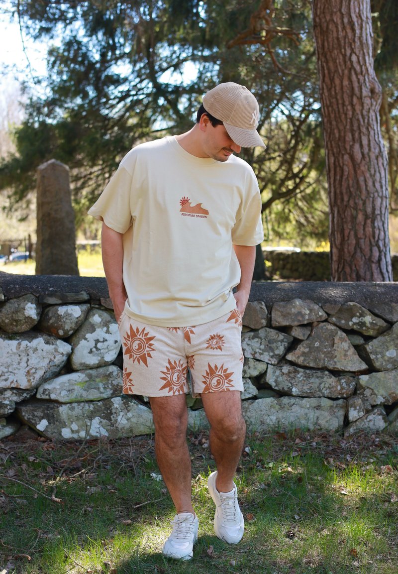 Homme avec casquette beige, t-shirt crème avec logo soleil, et shorts à motifs, debout sur l'herbe près d'un mur en pierre et d'arbres, regardant vers le bas, les mains dans les poches.