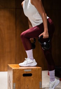 Kettlebell neri sono tenuti da una persona in scarpe da ginnastica bianche, leggings bordeaux e una canotta bianca, in piedi su una scatola di legno.