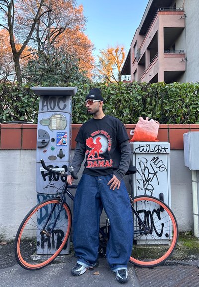 Hombre con gorro negro, gafas de sol, camiseta gráfica y vaqueros anchos, de pie junto a una bicicleta negra con llantas naranjas cerca de un parquímetro cubierto de grafitis y arbustos.