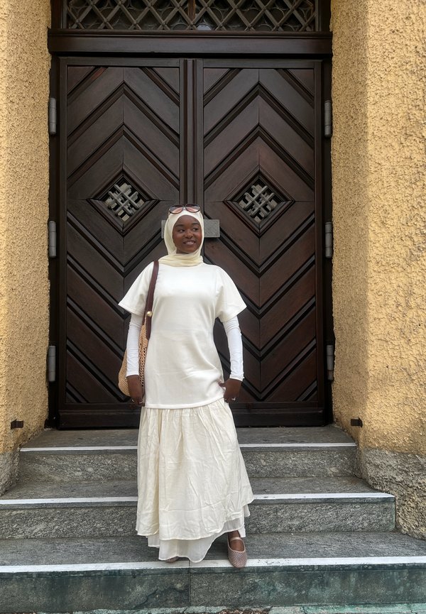 Mujer que lleva un hiyab blanco, un vestido blanco largo y gafas de sol está de pie en unos escalones de piedra frente a una gran puerta de madera oscura con patrones geométricos.