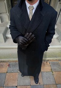 Man wearing a dark overcoat, check tie, black leather gloves, and black shoes standing on tiled steps outdoors.