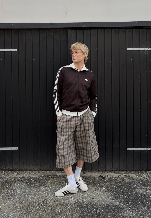 Osoba v čiernej bunde Adidas, károvaných širokých kraťasoch, bielych ponožkách a teniskách stojí s rukami v vreckách pred čiernymi drevenými dverami.