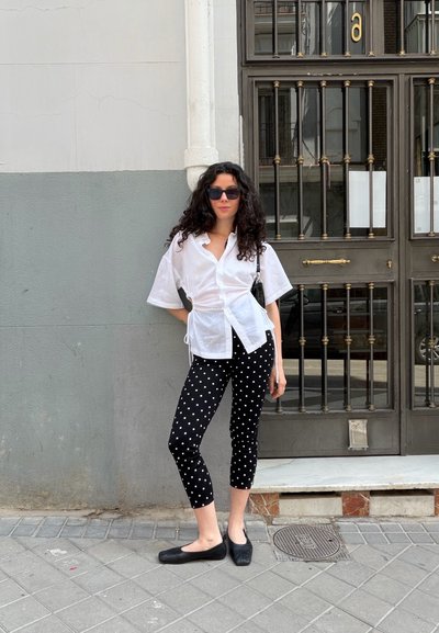 Mujer con pelo rizado y gafas de sol se apoya en una pared gris vistiendo una blusa blanca, pantalones negros con lunares y bailarinas negras en la acera de la ciudad.