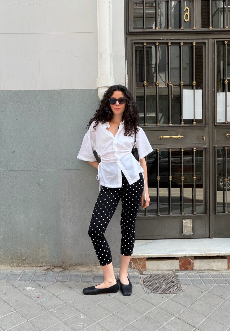 Femme aux cheveux bouclés et lunettes de soleil, appuyée contre un mur gris, vêtue d'un chemisier blanc, d'un pantalon noir à pois et de ballerines noires sur le trottoir en ville.
