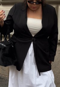 Blazer noir sur mesure avec une taille cintrée et une boucle en métal, porté sur un haut blanc et une jupe blanche fluide ; tenant un sac à main noir.