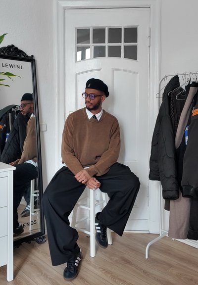 Suéter marrón de punto con un cuello blanco, pantalones negros oversize, boina negra y gafas. Reflexión en un espejo, suelo de madera beige.