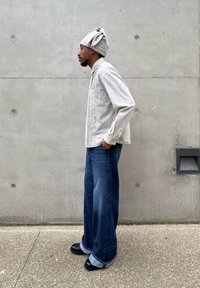 Homme debout de profil, portant une chemise à carreaux grise, un jean bleu à jambes larges, des chaussures noires et un turban gris, devant un mur en béton.