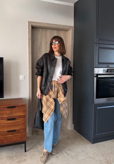 Mujer de pie en el interior con chaqueta de cuero negra, camisa blanca, vaqueros, gafas de sol, camisa de cuadros atada a la cintura, sosteniendo un bolso negro.