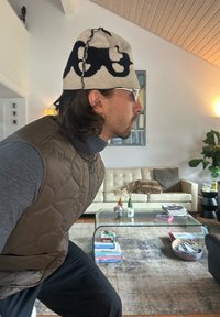 Strikket beige lue med svart abstrakt design, brun quiltet vest, grå turtleneck, klare briller, i et lyst interiør med sofa og glassbord.