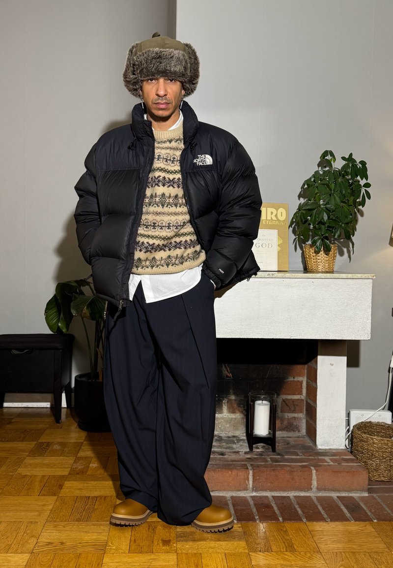 Homme portant un chapeau en fourrure, une doudoune noire, un pull à motifs, une chemise blanche, un pantalon large bleu marine et des bottes beige, debout à l'intérieur près d'une cheminée blanche avec des plantes.