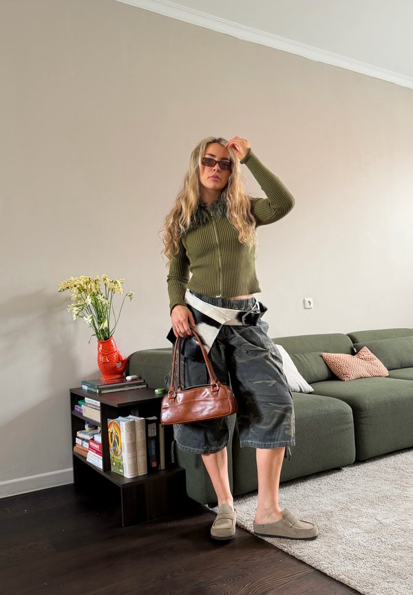 Persoană cu păr blond lung, purtând ochelari de soare, pulover verde, pantaloni largi camuflați, ținând o geantă maro, stând lângă o canapea verde în sufragerie.