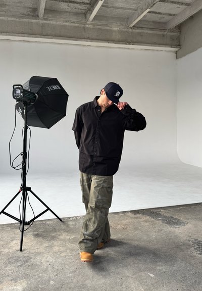 Camisa negra de botones, pantalones cargo oliva y botas de trabajo color beige. Configuración de estudio con un soporte de luz y fondo de softbox.
