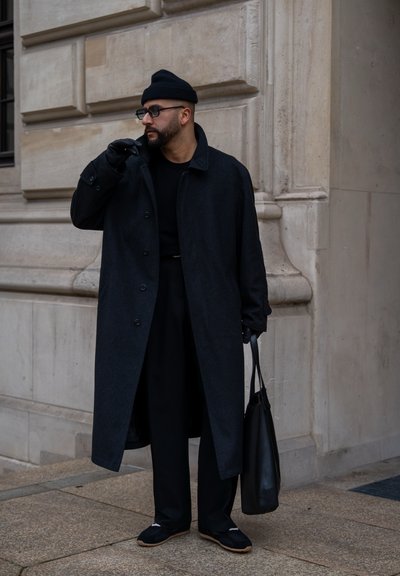 Hombre con abrigo negro, gorro, guantes y gafas, sosteniendo una bolsa tote negra, de pie contra una pared de piedra.