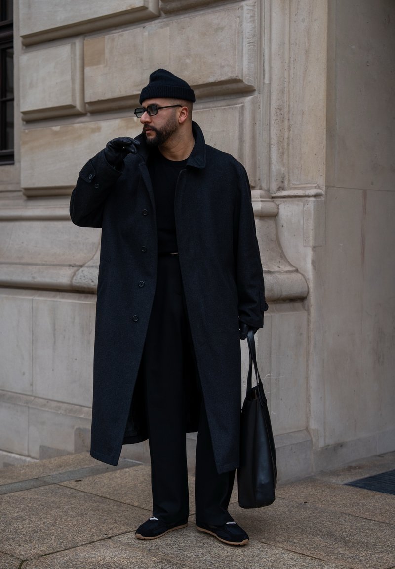 Άνδρας με μαύρο παλτό, σκούφο, γάντια και γυαλιά, κρατώντας μια μαύρη τσάντα ώμου, στέκεται μπροστά σε τοίχο πέτρινης οικοδομής.