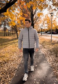 Γκρι φουσκωτό μπουφάν με κουκούλα, με οριζόντιο σχέδιο καπιτονέ; φορεμένο με σκούρα τζιν και λευκά sneakers; φθινοπωρινά φύλλα στο βάθος.