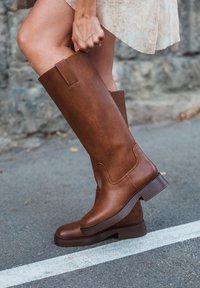 Brune skinnknehøye støvler med en glatt tekstur, rund tå og solid blokkheel; har en sideløs pull-tab og kontraststing.